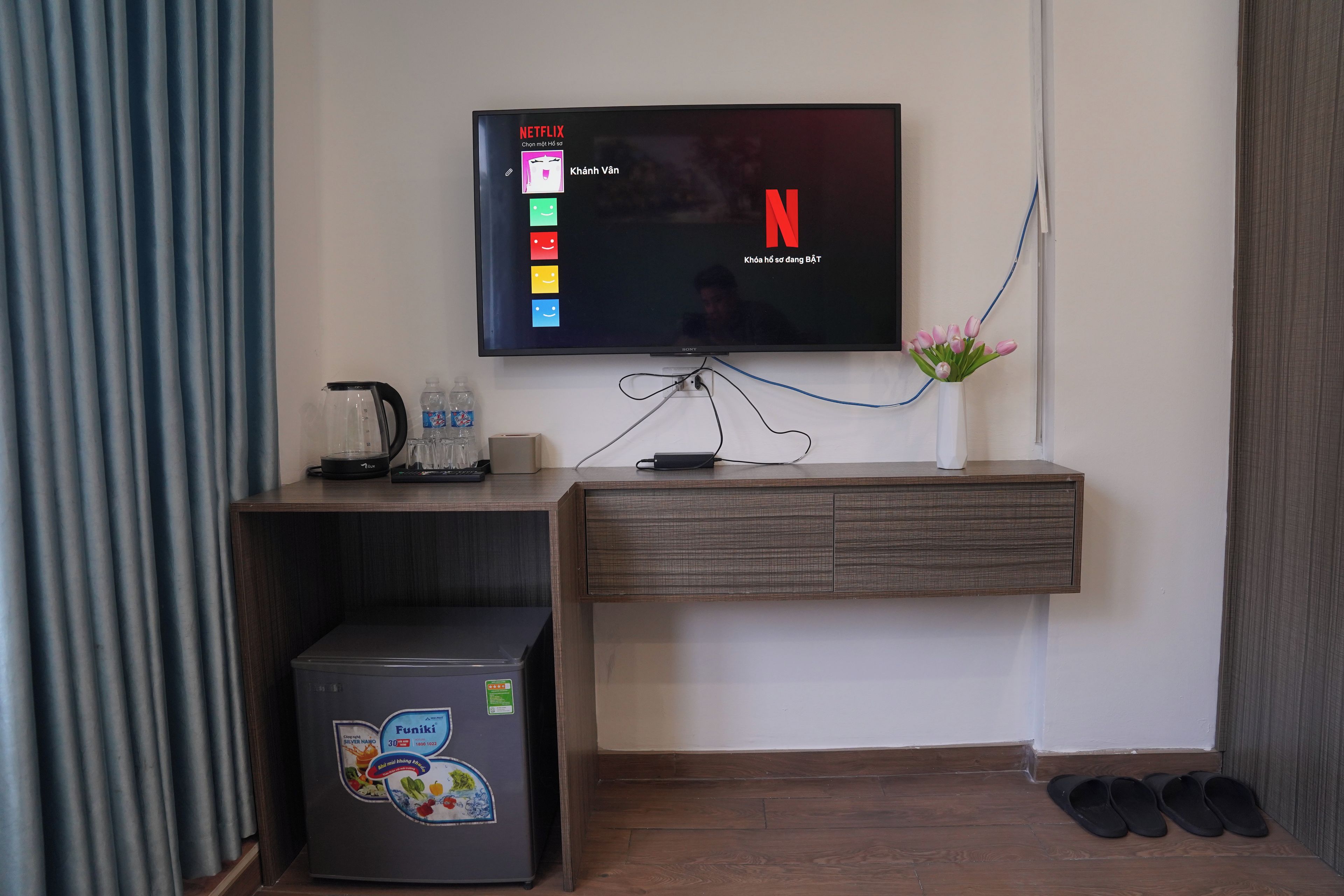 Una sala moderna con un televisor de pantalla plana montado en la pared, una cafetera, una botella de agua, un refrigerador pequeño y un jarrón con flores.