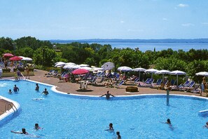 Ein Pool mit mehreren Liegestühlen und Sonnenschirmen, umgeben von Grünflächen und Blick aufs Meer.