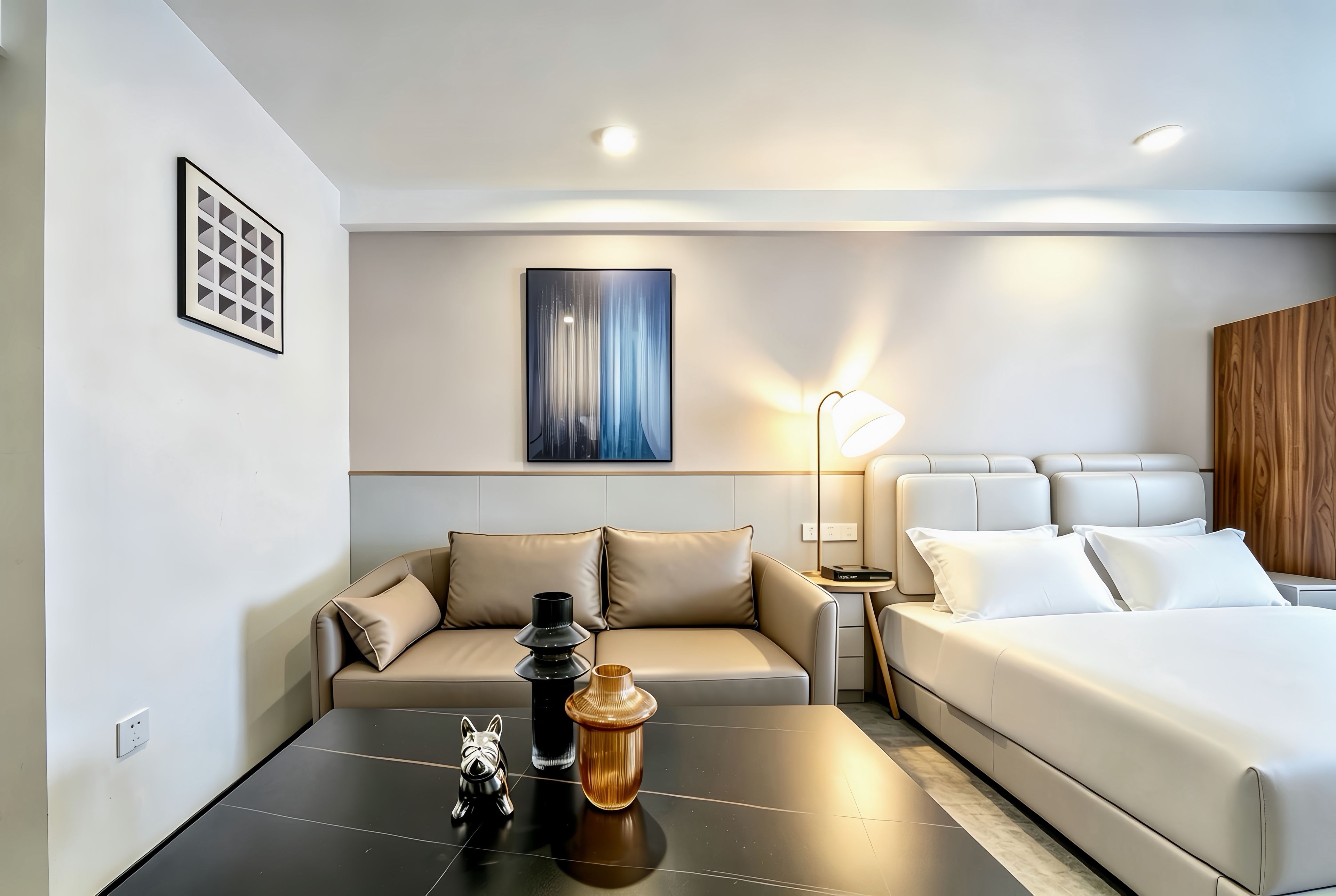 A modern hotel room with a sofa, a bed, a small table with decorative items, and a wall-mounted artwork.
