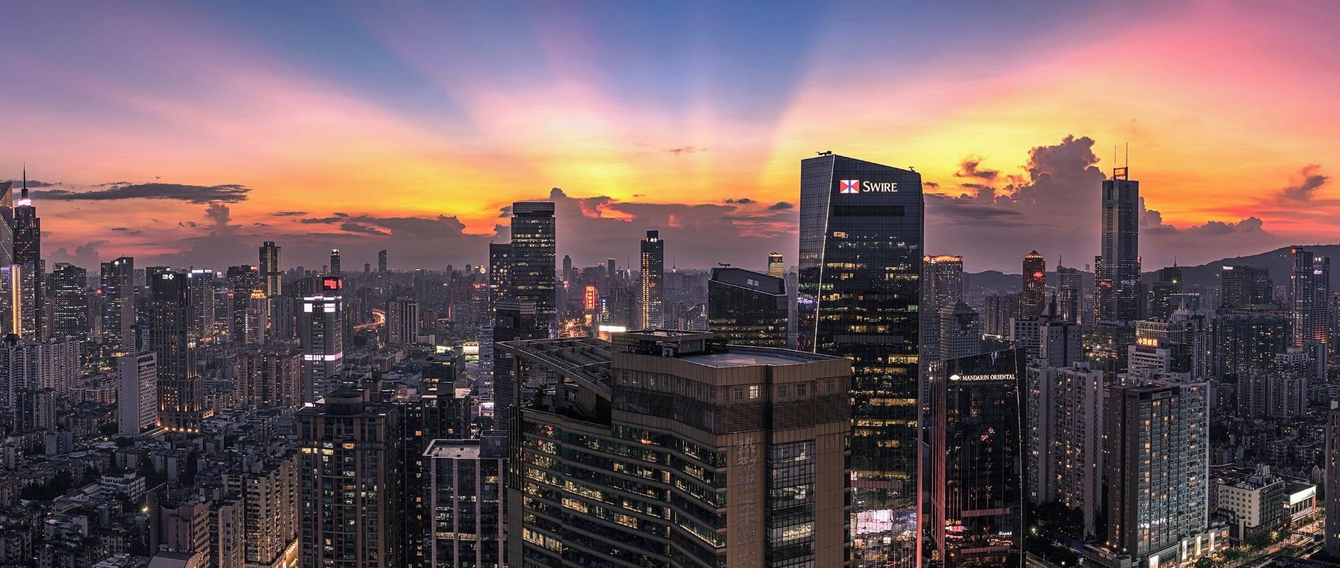 A cityscape at dusk with illuminated skyscrapers and a prominent SWIRE building.