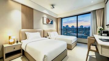 A hotel room with two beds, a desk, a chair, a large window with a city view, and a ceiling-mounted projector.