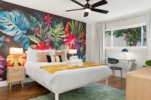 Une chambre d’hôtel comprenant un lit, des tables de chevet, un bureau, une chaise, un ventilateur de plafond et une grande fresque murale aux motifs tropicaux.