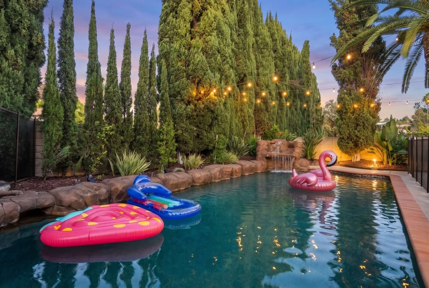 A swimming pool with inflatable toys, surrounded by tall trees and a waterfall feature.