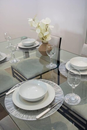 Een eettafel gedekt met witte borden, bestek en wijnglazen, met een vaas witte bloemen.