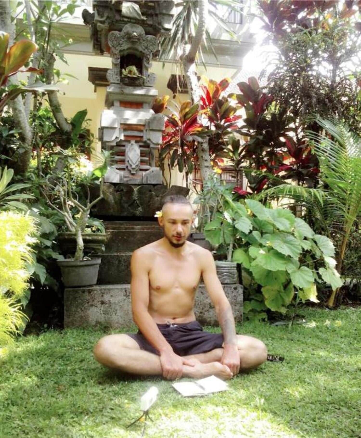 A person sitting on the grass in a garden with various plants and a stone structure in the background.