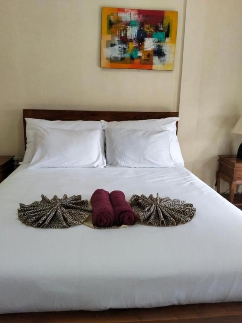 A hotel room with a large bed, two pillows, a colorful abstract painting on the wall, and a neatly arranged towel and patterned cloth on the bed.