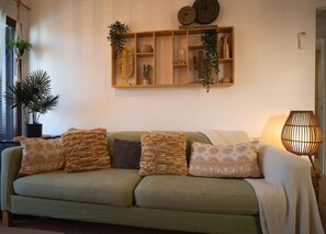 Un sofà verd amb coixins estampats, una estant de paret amb objectes decoratius i una làmpada de peu.