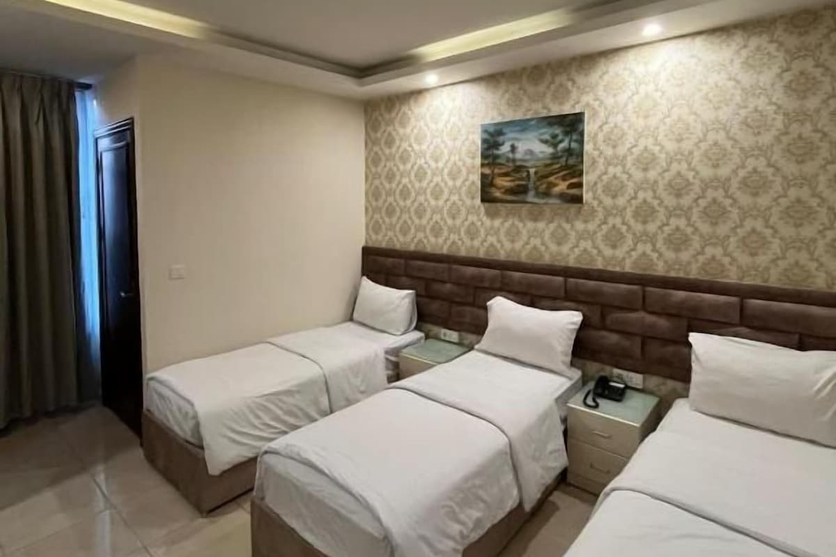 Habitación de hotel con tres camas, una mesita de noche, un cuadro en la pared y papel tapiz estampado.