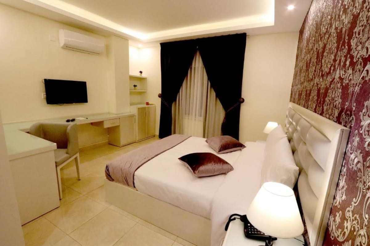 Una habitación de hotel con cama, mesita de noche, lámpara y televisión en la pared.