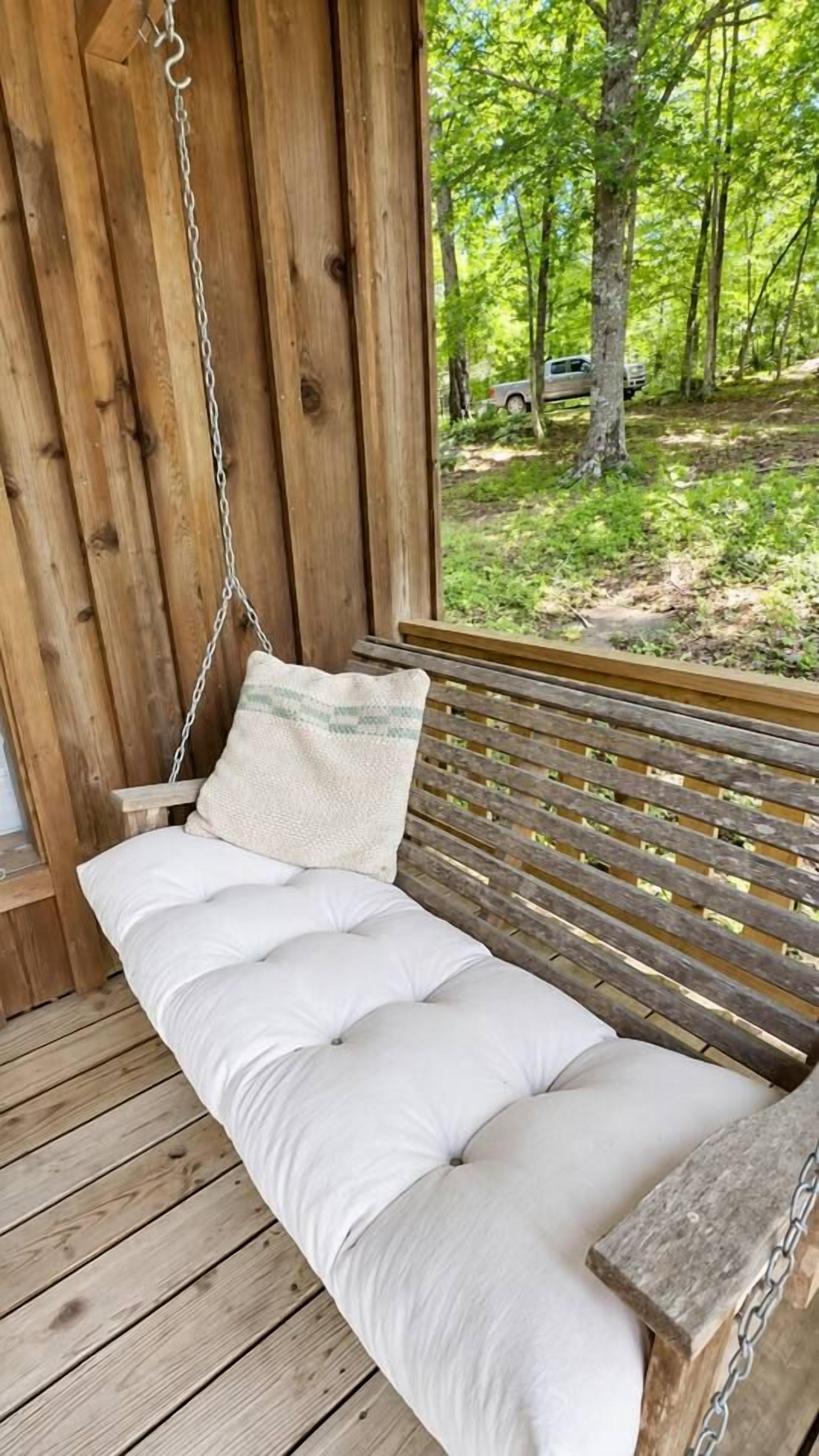 A wooden swing bench with cushions, a chain, and a view of a forested area.