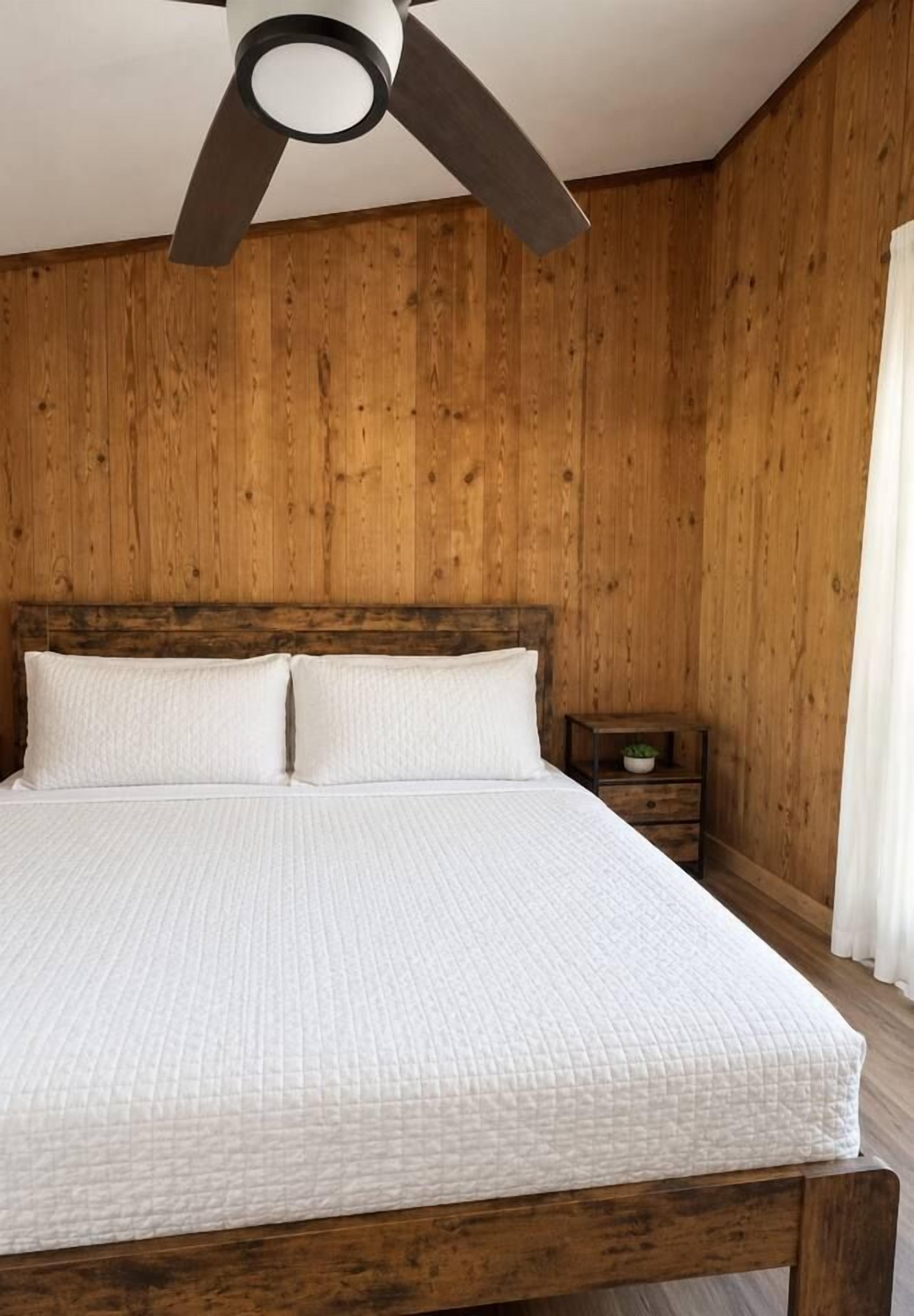 Une chambre à coucher avec une tête de lit en bois, un lit avec une literie blanche, une table de chevet avec une plante et un ventilateur de plafond.