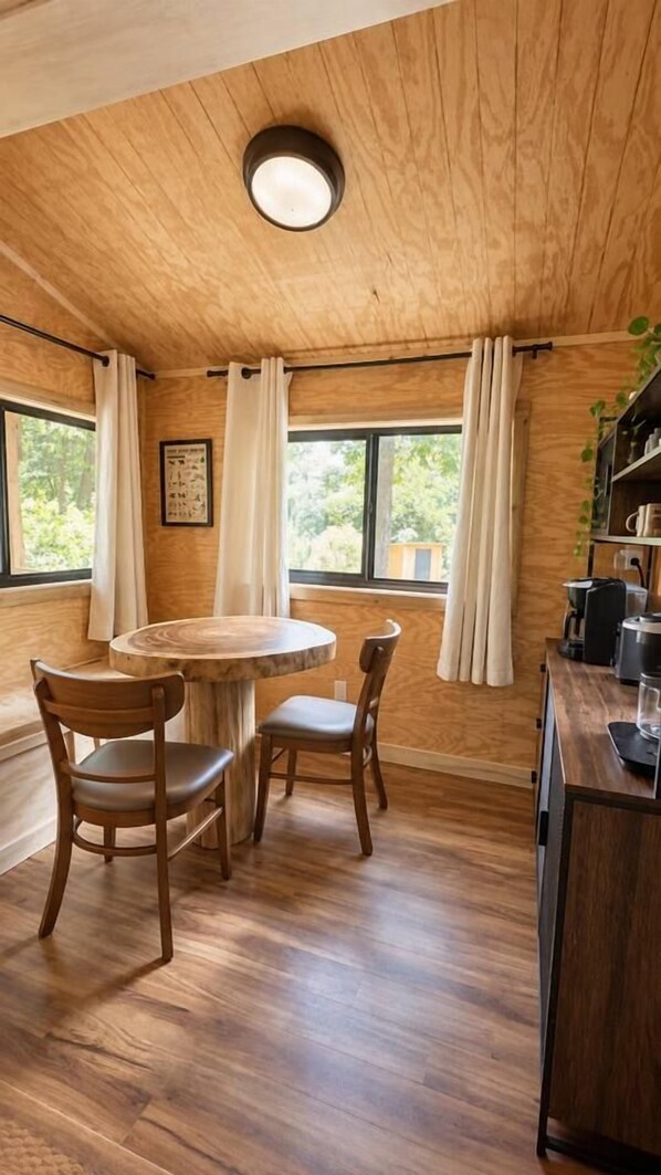 A wooden interior with a round table and chairs, a ceiling light, and windows with curtains.