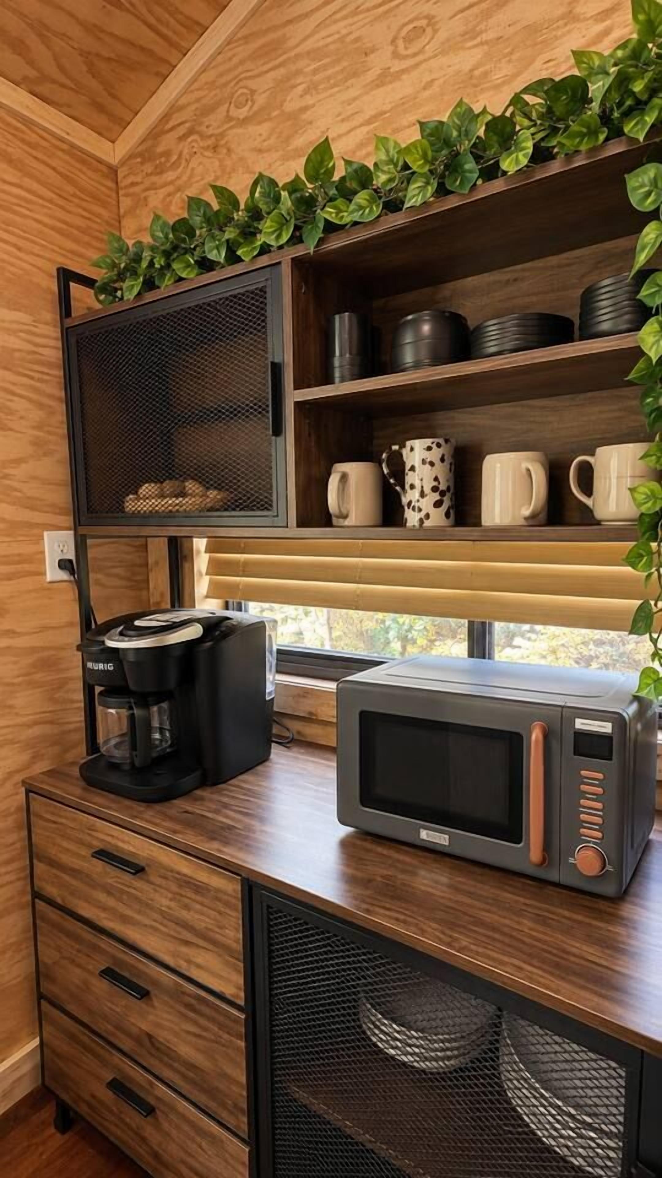 A kitchenette with a coffee maker, microwave, and shelves with mugs and a plant.