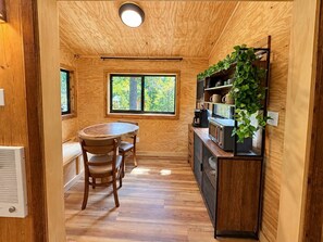 A compact kitchen with a dining table and chairs, a built-in shelf with plants, and a window providing natural light.