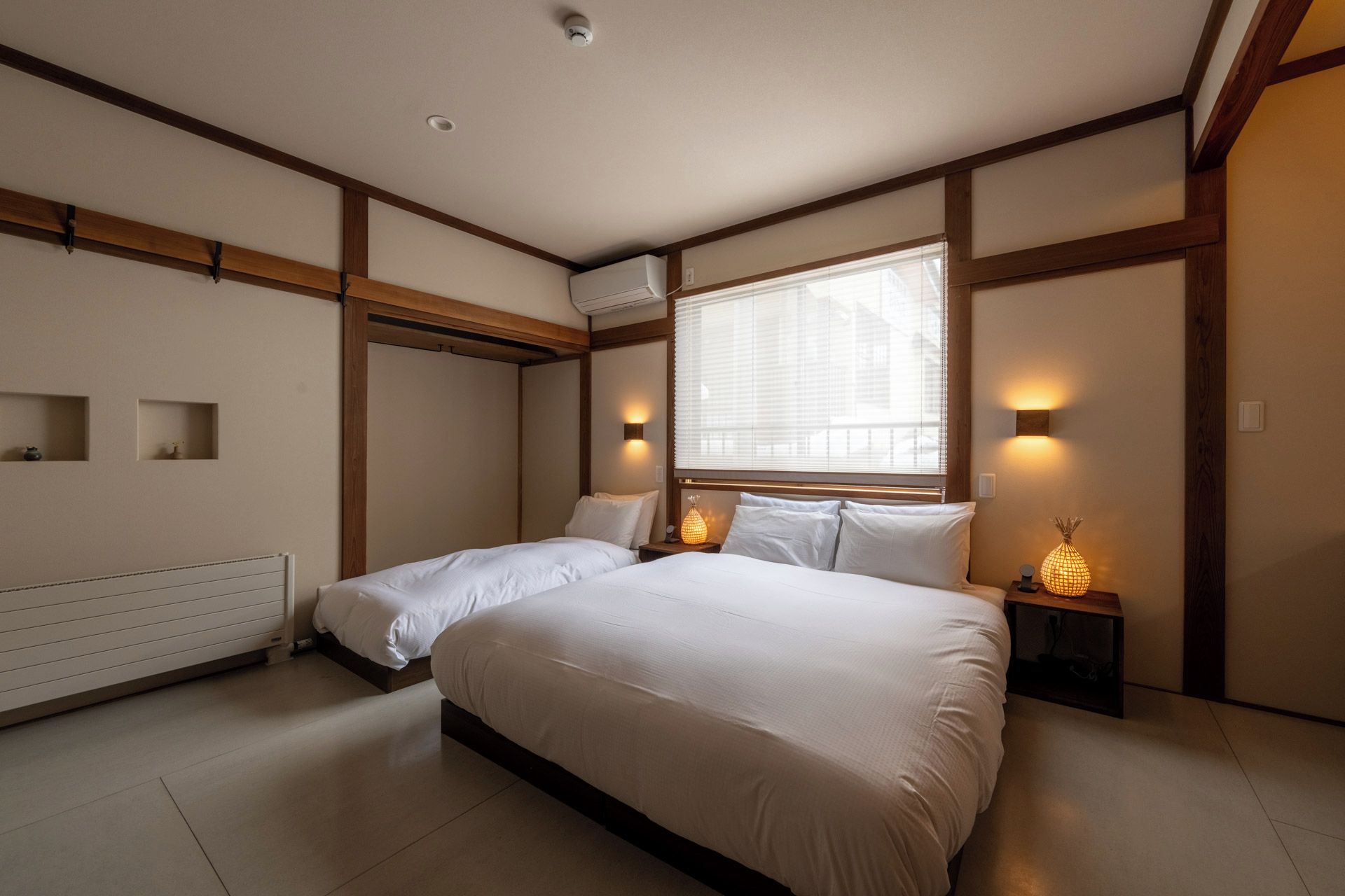A bedroom with a large bed, two bedside tables with lamps, a radiator, and a window with blinds.