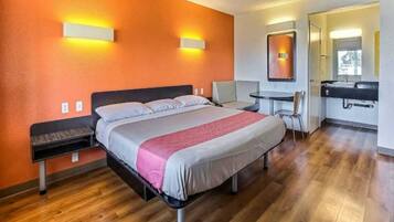 A hotel room with a bed, bedside tables, a desk, a chair, and a bathroom with a mirror and sink.