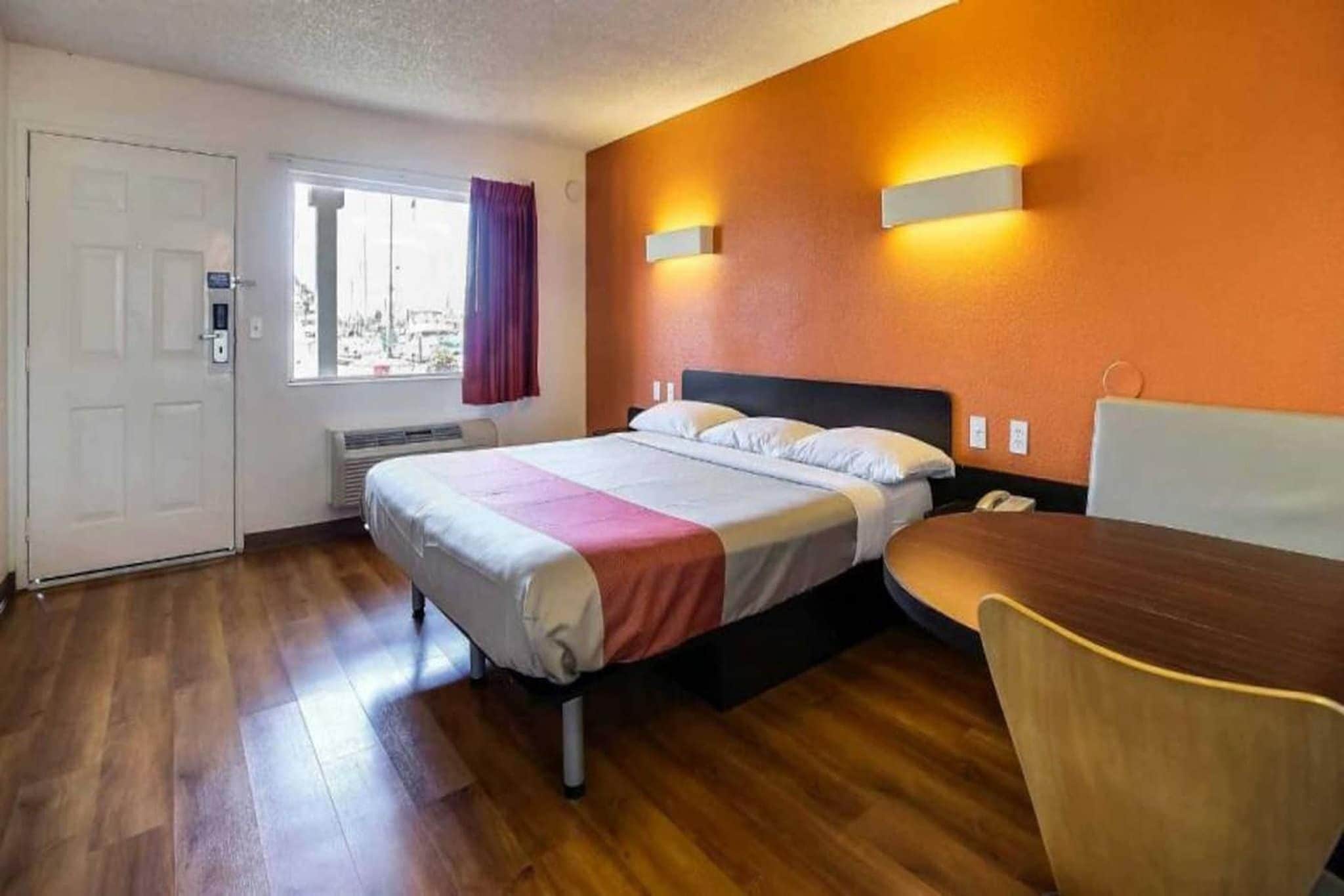 A hotel room with a bed, a desk, and a chair. There is a window with curtains, a door, and a wall-mounted light fixture.