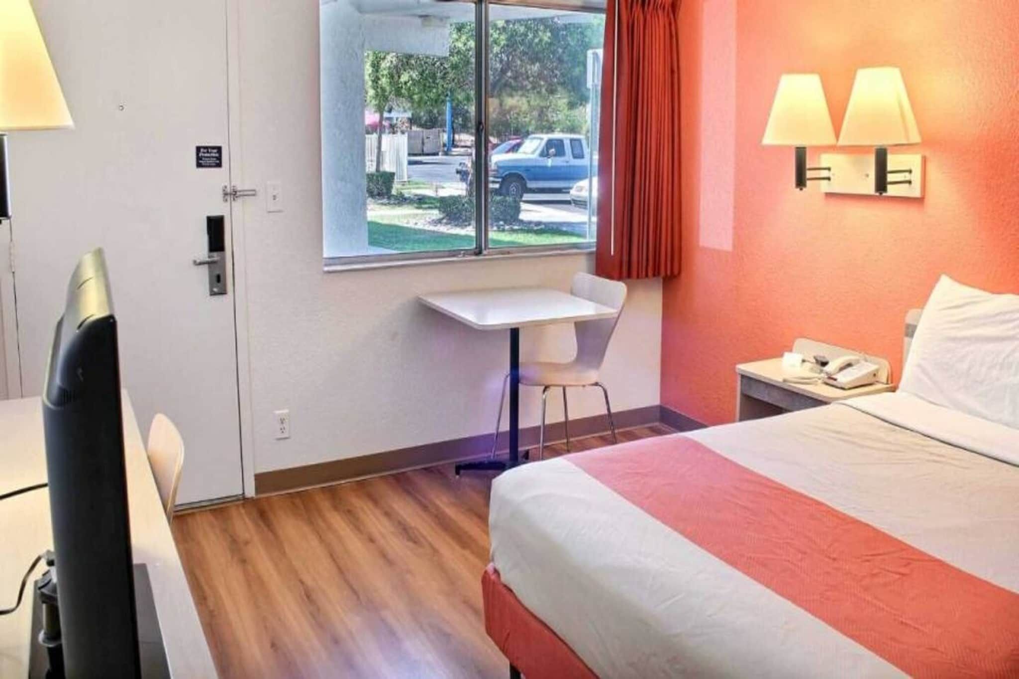 Quarto de hotel com cama, escrivaninha, cadeira, janela com vista para a rua e telefone na mesa de cabeceira.