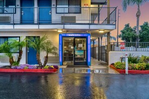 A hotel entrance with a blue 'welcome' sign, a gated driveway, and a palm tree-lined pathway. - 2 Full Beds | Mini Fridge (El Monte)