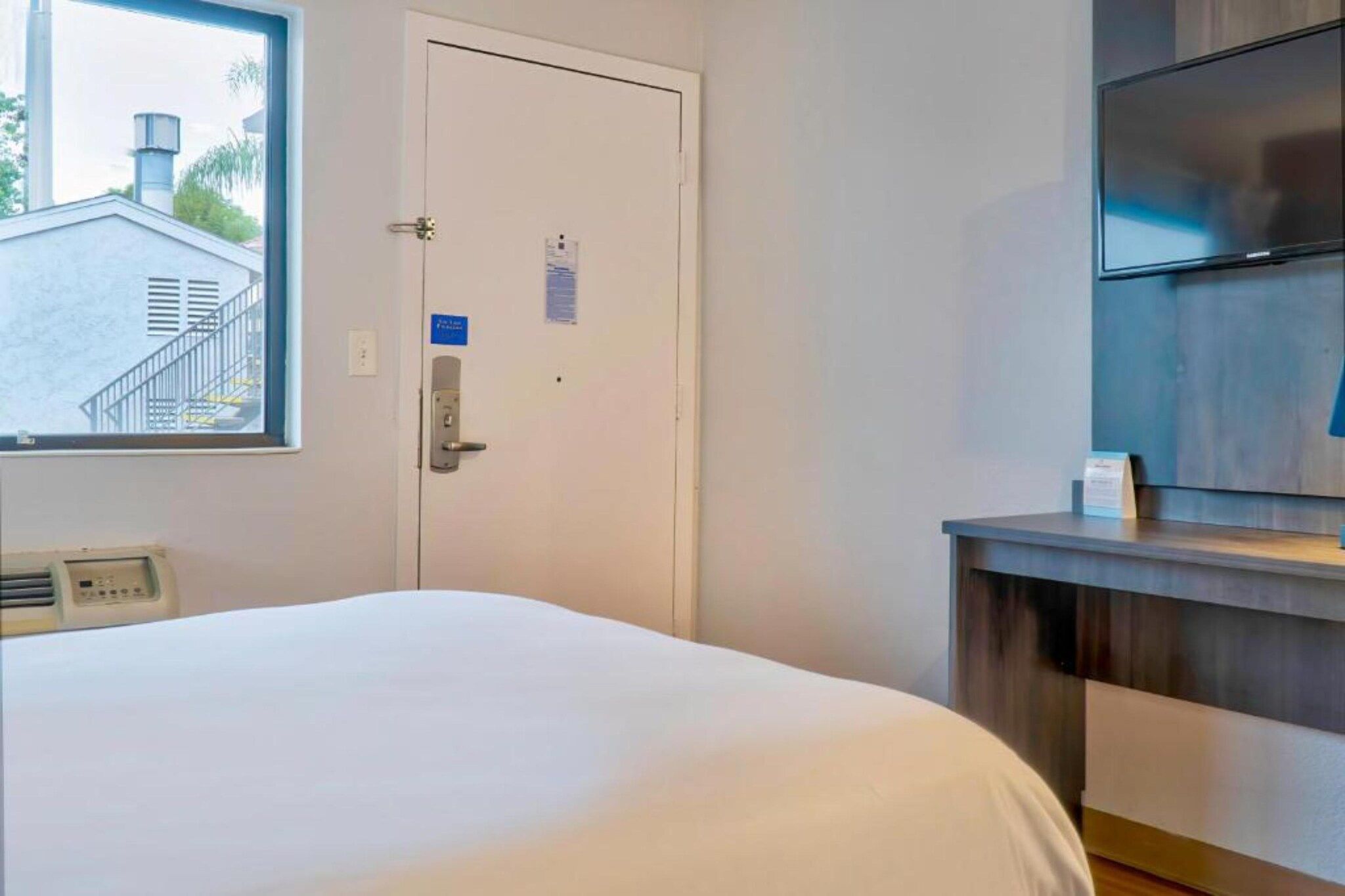A hotel room with a bed, a door with a lock, a window with a view of a building, and a flat-screen TV on a wooden stand.