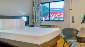 A hotel room with a large bed, a desk, a chair, a window with a view of a Coca-Cola sign, and a patterned curtain.