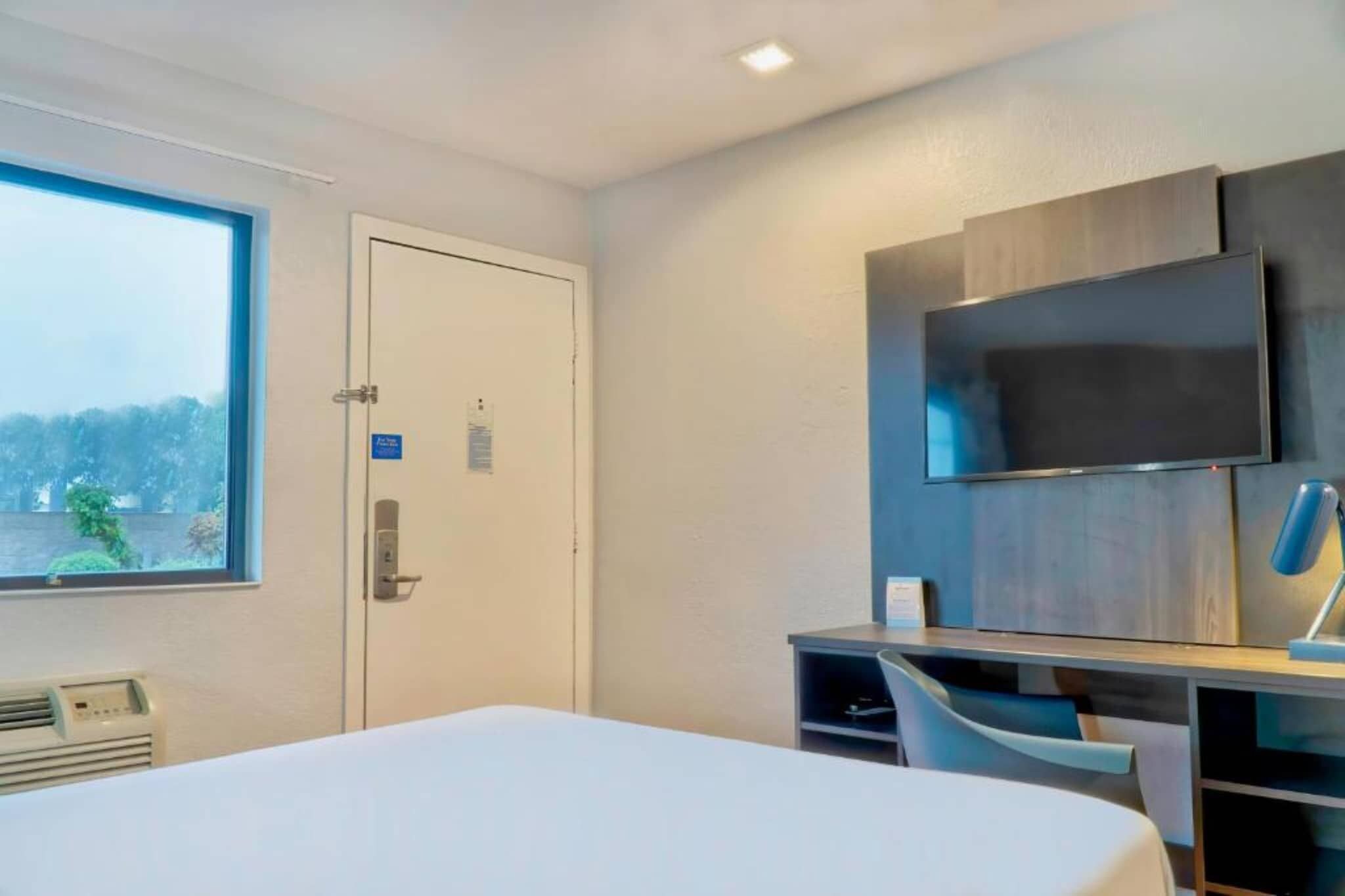 Una habitación de hotel con cama, escritorio con silla, televisor montado en la pared y ventana con vista a árboles.
