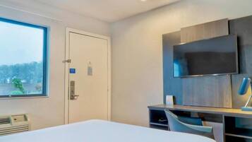Una habitación de hotel con cama, escritorio con silla, televisor montado en la pared y ventana con vista a árboles.