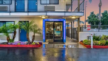 A hotel entrance with a blue 'welcome' sign, a gated driveway, and a palm tree-lined pathway.