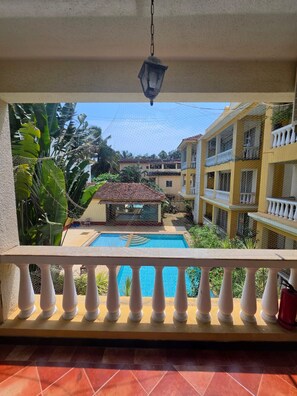 A balcony with a view of a swimming pool, surrounded by lush greenery and a building with balconies. - Charming 2-bedroom apartment with AC , POOL & GYM in gorgeous Goa (Goa)
