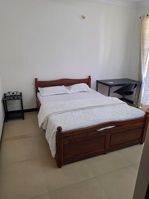 A single bed with a wooden frame and white bedding, a small bedside table, a desk with a chair, and a curtain on the window. - Charming 2-bedroom apartment with AC , POOL & GYM in gorgeous Goa (Goa)