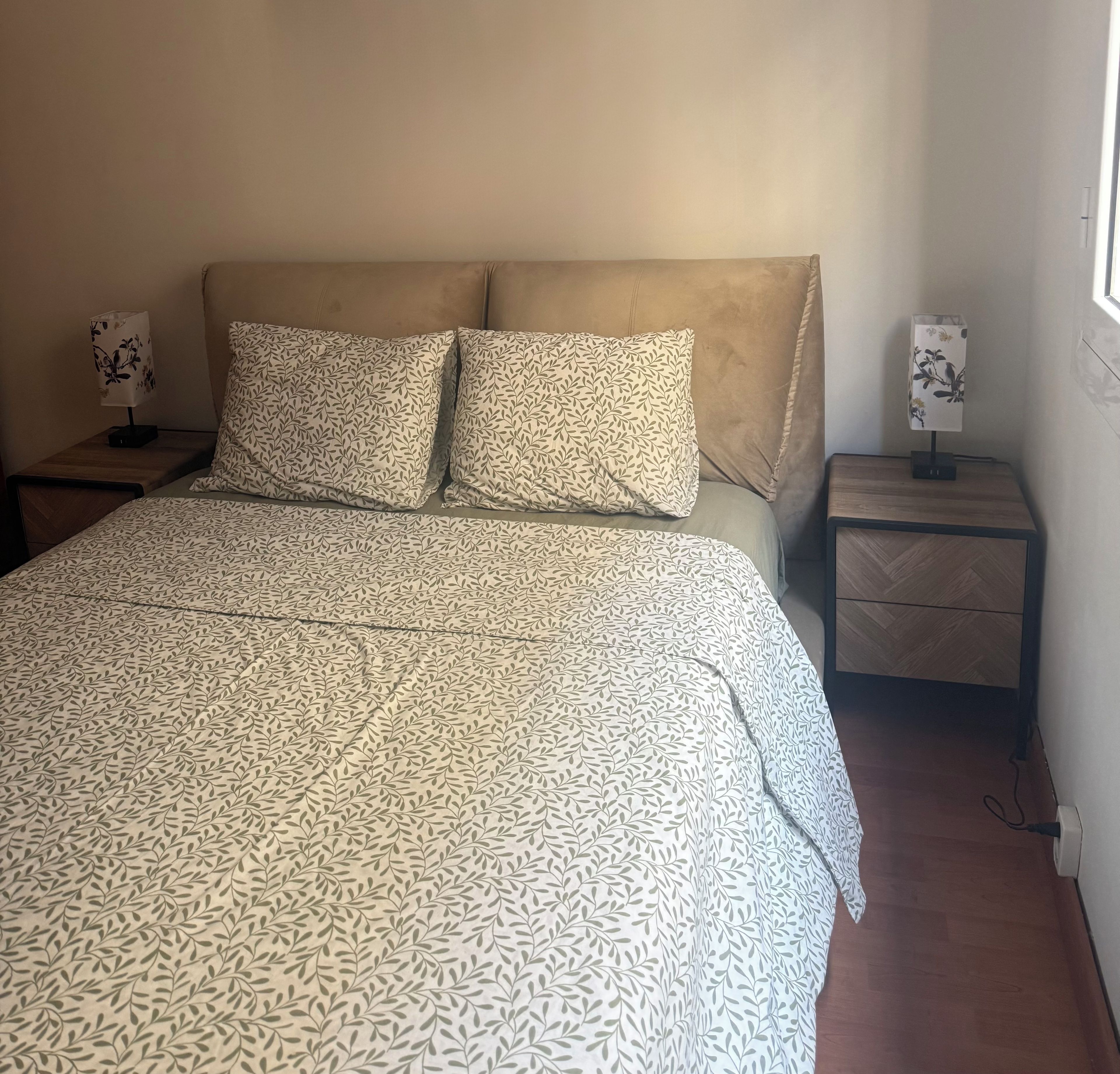 Una cama con una colcha estampada, dos almohadas, una mesita de noche con una lámpara y suelo de madera.