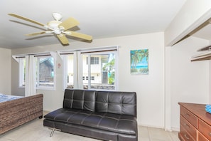 A room with a black leather sofa, a ceiling fan, and a window with a view of a building and greenery. - Monthly Stays! Waimanalo Beach Cottages, Unit 1 (Waimanalo)