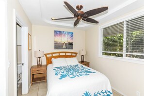Ein Schlafzimmer mit einem Bett, einem Deckenventilator, einem Fenster mit Jalousien, einem Nachttisch und einer an der Wand befestigten Lampe.