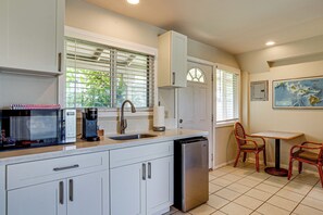 A kitchen with white cabinets, a stainless steel dishwasher, a coffee maker, and a window with blinds. - Walk to Shore! Waimanalo Beach Cottages, Unit 9 (Waimanalo)