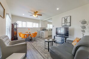 Un salon moderne avec un canapé, une table basse et un téléviseur. On y trouve également un coin repas avec des chaises orange et une table. La pièce est équipée d’un ventilateur de plafond et d’une grande fenêtre avec des stores.