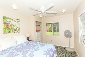 Ein Schlafzimmer mit einem Bett, einem Deckenventilator, einem Fenster mit Jalousien, einem Standventilator und einer Tür zu einem Schrank.