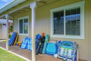 Uma casa com varanda, janela com persianas e diversos objetos externos, como uma prancha de surf azul e amarela, uma bolsa listrada em azul e branco, e um carrinho de metal com uma sacola azul e uma bola de futebol.