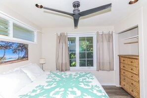 A bedroom with a bed, a ceiling fan, a window with curtains, a dresser, and a painting on the wall. - Walk to Ocean! Waimanalo Beach Cottages: Unit 5 (Waimanalo)