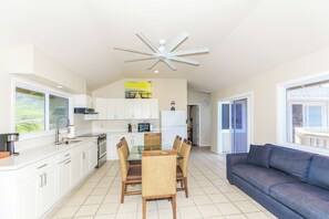 A modern kitchen with white cabinets, a dining table with chairs, a ceiling fan, and a view of the outdoors through a window. - Extended Escape! Waimanalo Beach Cottages, Unit 11 (Waimanalo)