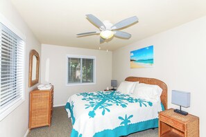 A bedroom with a bed, a ceiling fan, a window with blinds, a bedside table with a lamp, and a painting on the wall. - Steps to Beach! Waimanalo Beach Cottages, Unit 10 (Waimanalo)