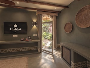 A modern living room with a television displaying the Khubani Beach Resort logo, a fireplace, a large window with curtains, and a woven basket on the wall. - Khubani Resort (Ashvem)