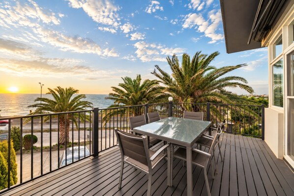 A balcony with a table and chairs, overlooking a road and the sea.