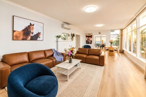 A spacious living room with a brown leather sofa, a blue armchair, and a coffee table. There is a painting of horses on the wall, a mirror, and a view of the ocean through large windows.