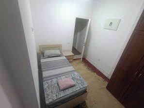Ein kleines, schmales Zimmer mit einem Einzelbett, einem Holzschrank und einem Spiegel an der Wand.