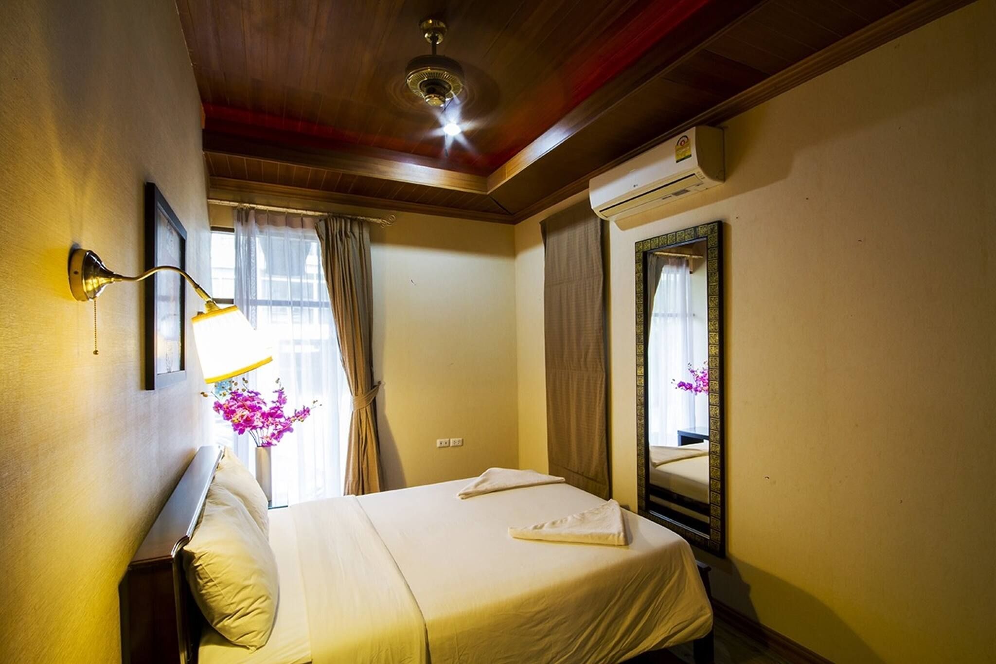 Une chambre d’hôtel comprenant un lit, une lampe fixée au mur, un appareil de climatisation, une fenêtre avec des rideaux et un miroir avec un cadre décoratif.