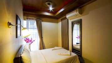 Une chambre d’hôtel comprenant un lit, une lampe fixée au mur, un appareil de climatisation, une fenêtre avec des rideaux et un miroir avec un cadre décoratif.