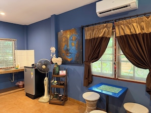 Una habitación con pared azul, un cuadro, una ventana con cortinas, un ventilador, una mesita y un refrigerador.