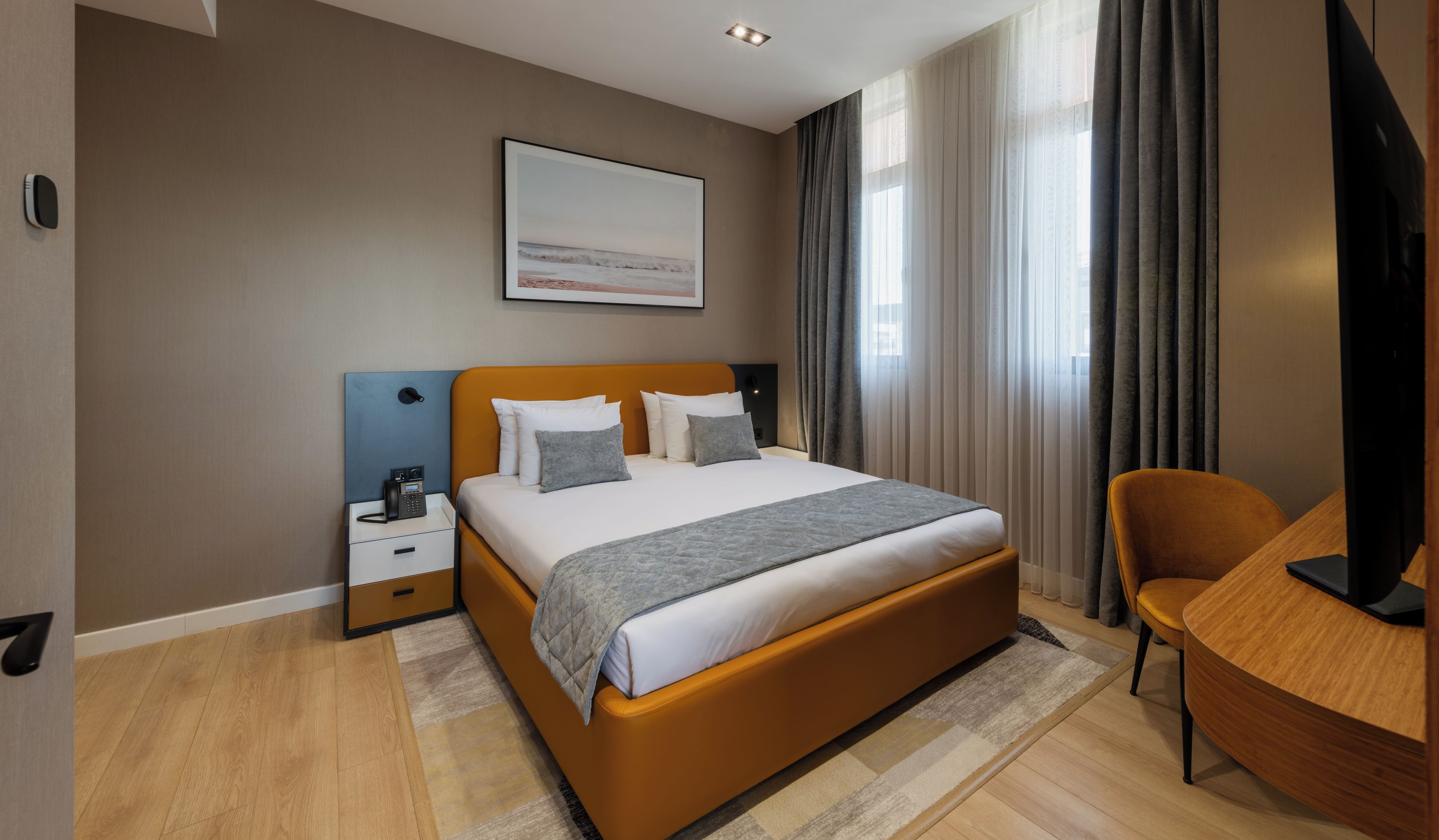 Una habitación de hotel moderna con cama, mesita de noche, silla y un ventanal con cortinas.