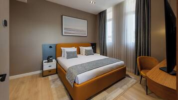 Una habitación de hotel moderna con cama, mesita de noche, silla y un ventanal con cortinas.
