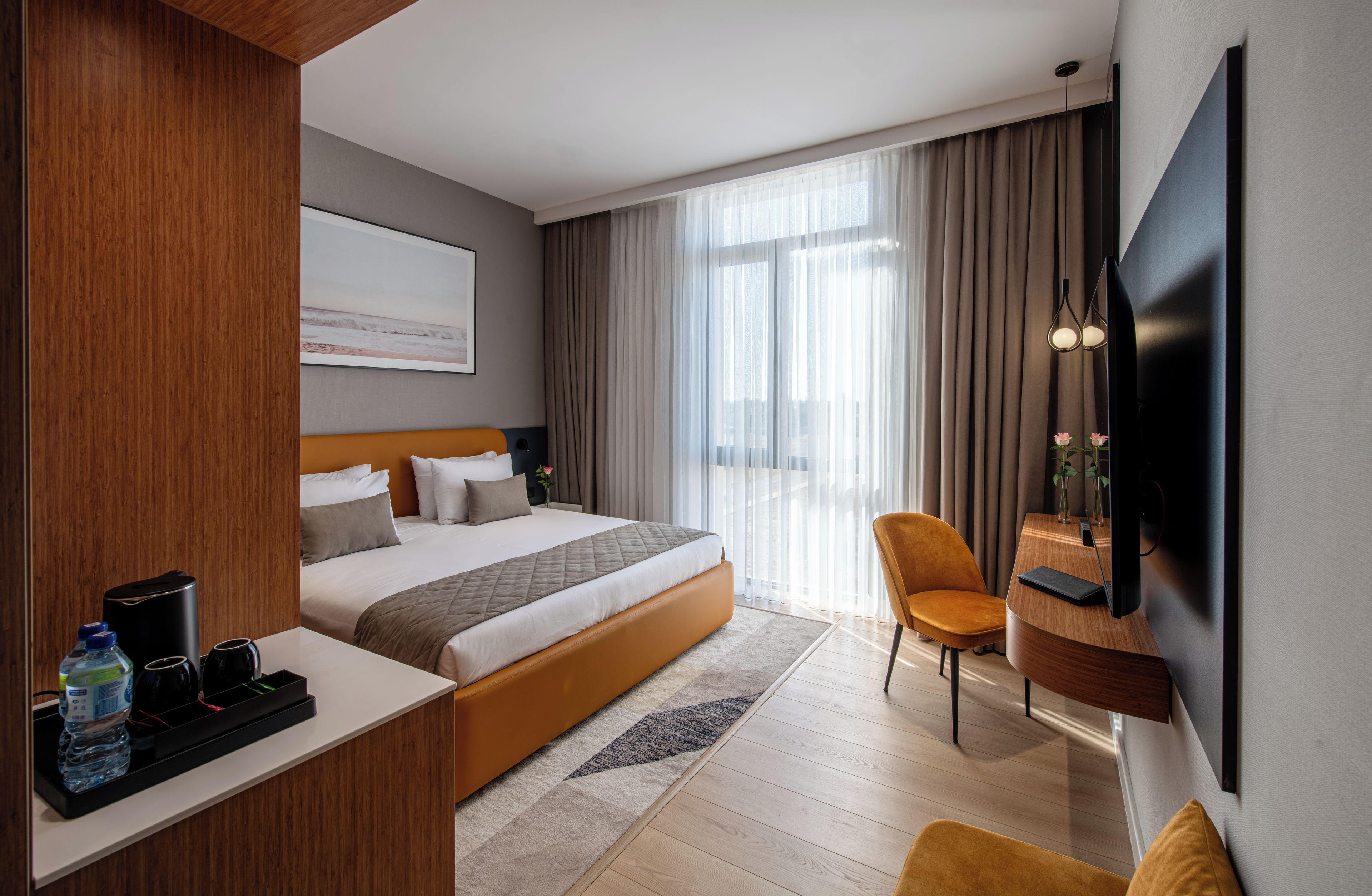 A hotel room with a bed, bedside table, chair, desk, TV, and a large window with curtains.